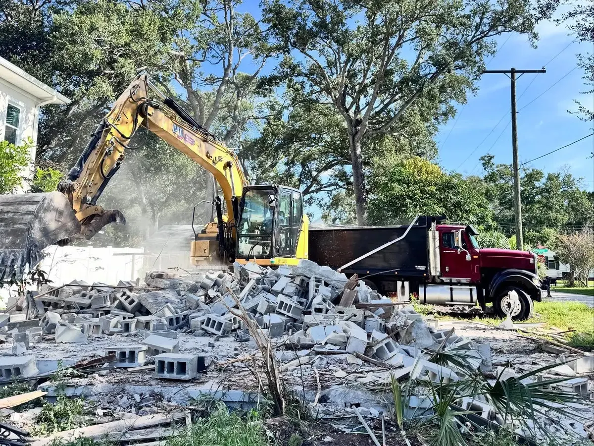 Excavator tearing down a concrete block house in Tampa, with a dump truck parked at the street